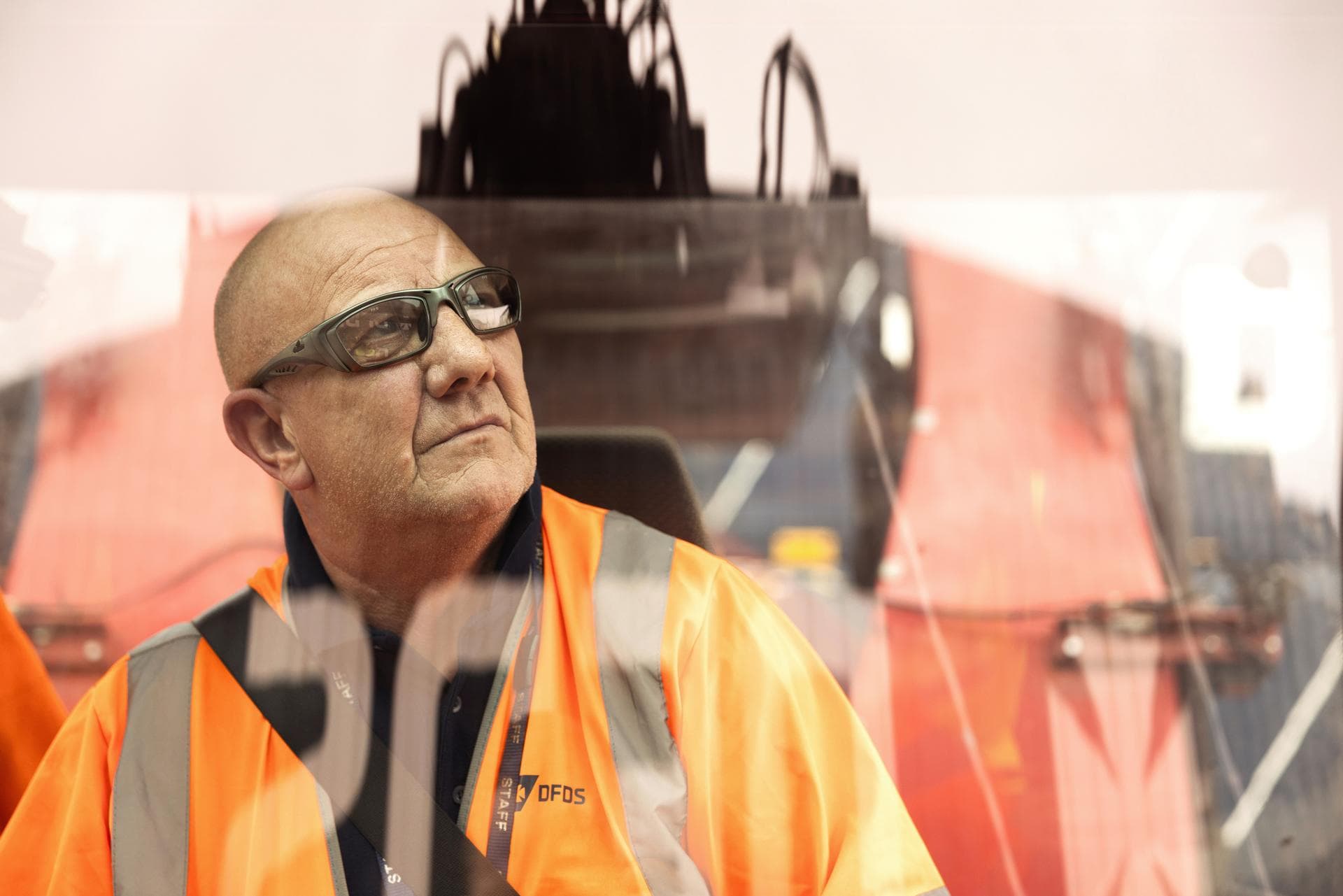 Crane driver inside a cabin, looking outside, wearing orange safety jacket, branded with DFDS