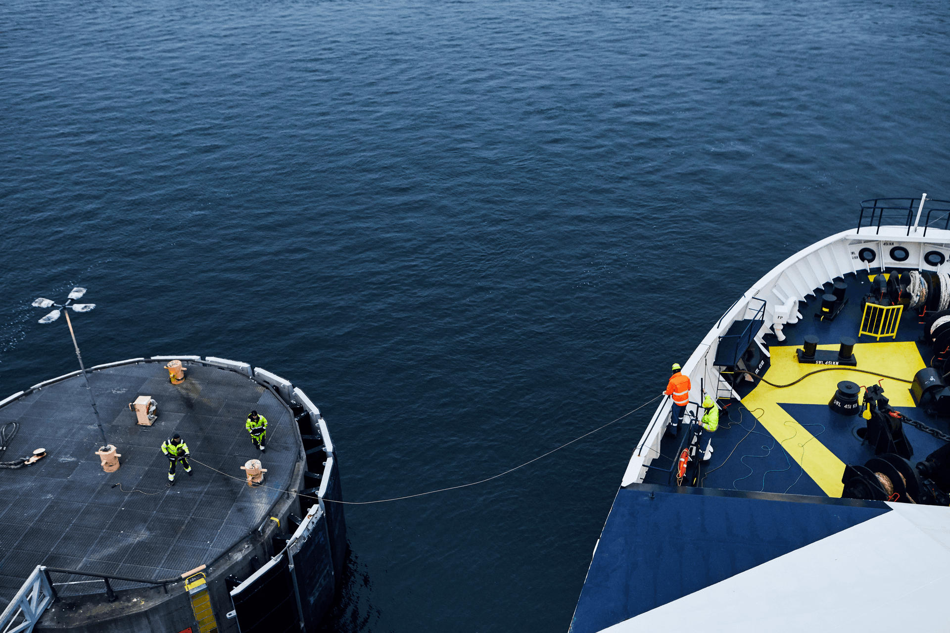 A DFDS vessel docks in the port: two seafarers with yellow security helmets on the deck working with the ropes and two people on the shore pulling in the ropes
