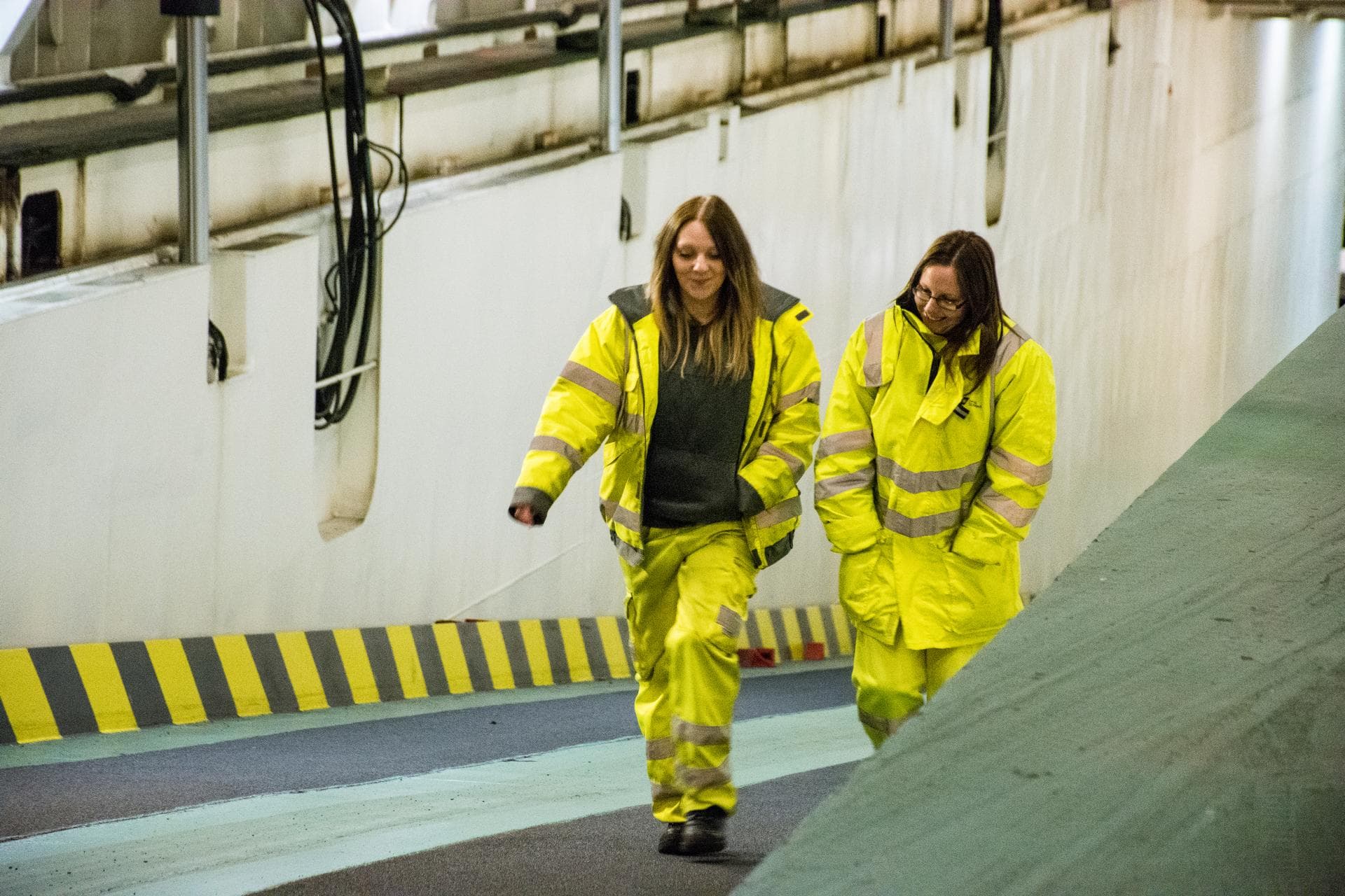 Two female ship workers, walking up on a ramp on a vessel, wearing yellow safety jackets, talking to each other