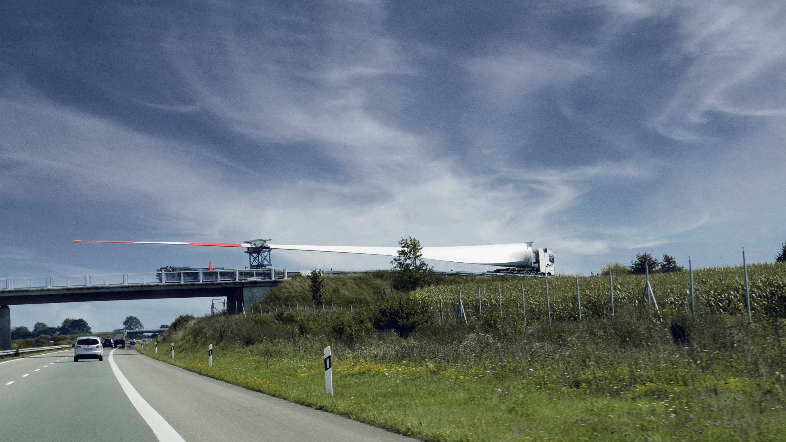 A wind turbine blade is transported by a DFDS truck crossing a bridge over a highway