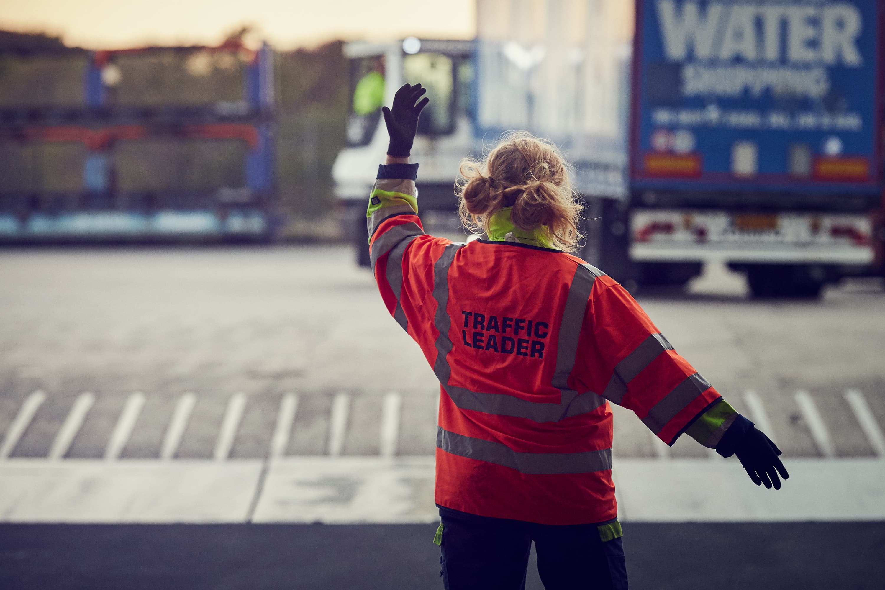 A woman wearing an orange vest that says Traffic Leader signals to a truck in the background
