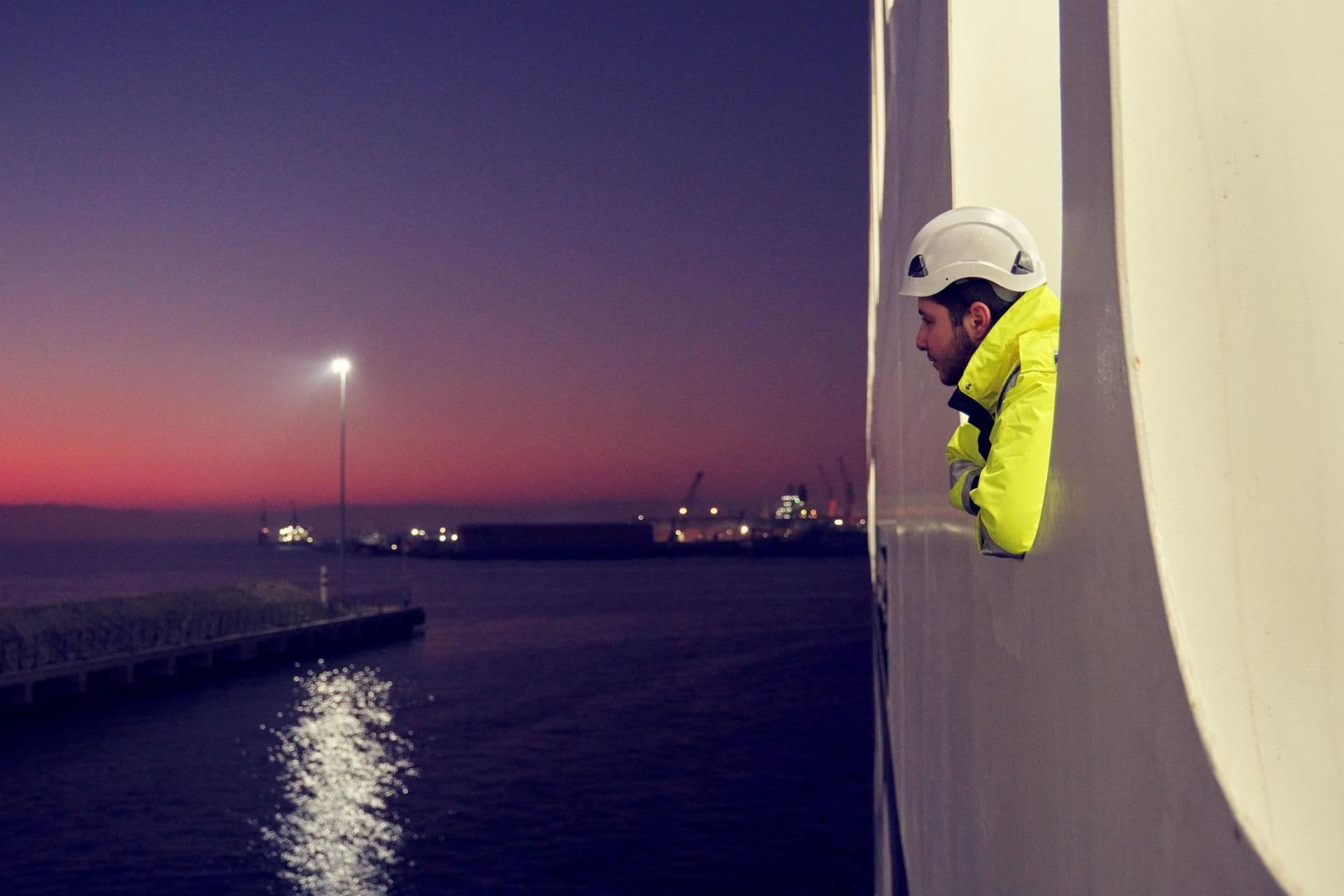 Man with yellow security jacket and white helmet, looking out of a porthole of a vessel, at sunset in harbour of Pendik/TR