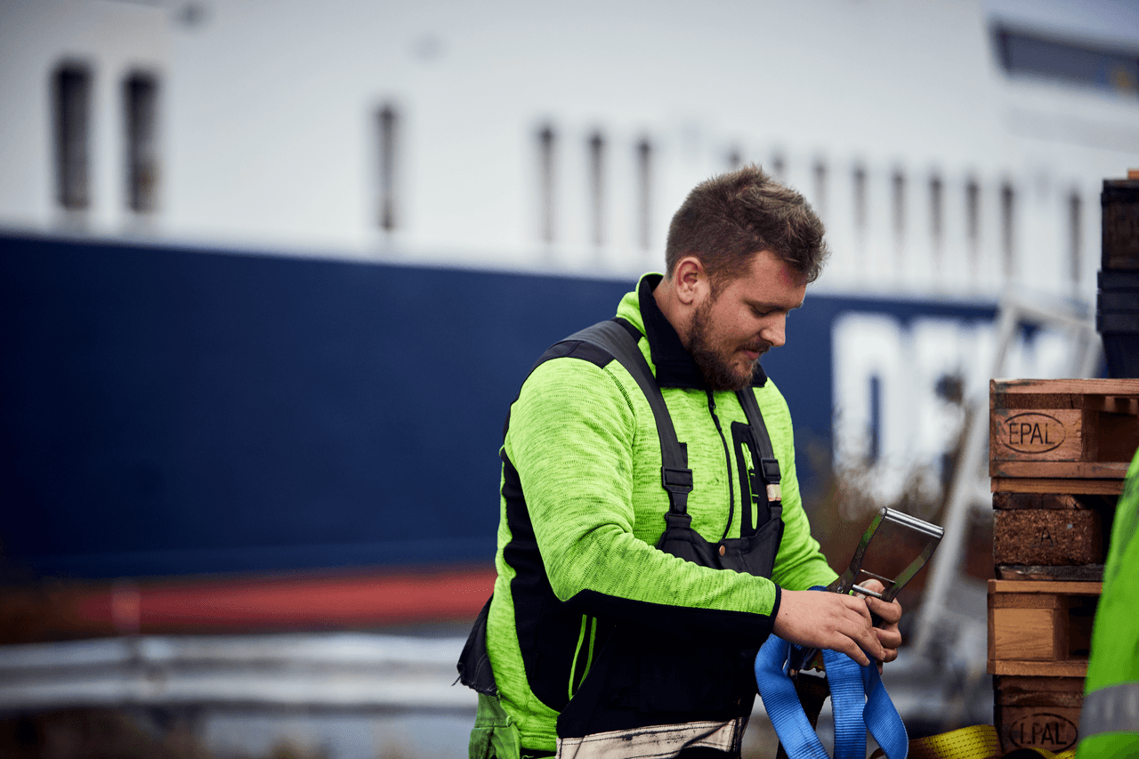 A terminal with a yellow security jacket, working in the harbour with wooden pallets, within the background, a DFDS vessel