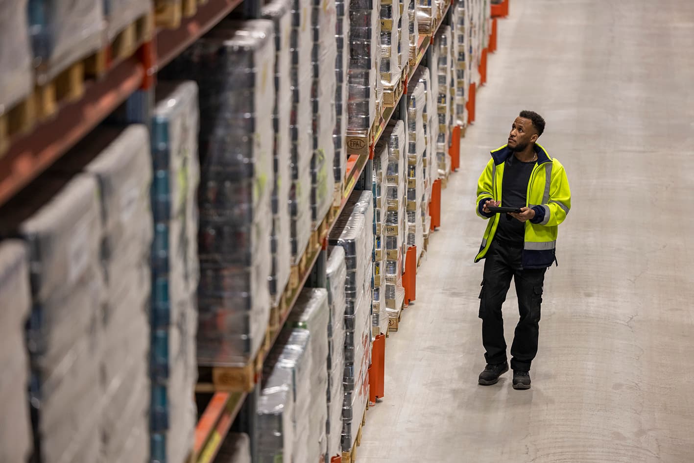 Man in yellow jacket, standing in the warehouse before loaded storage rack, with tablet in hand
