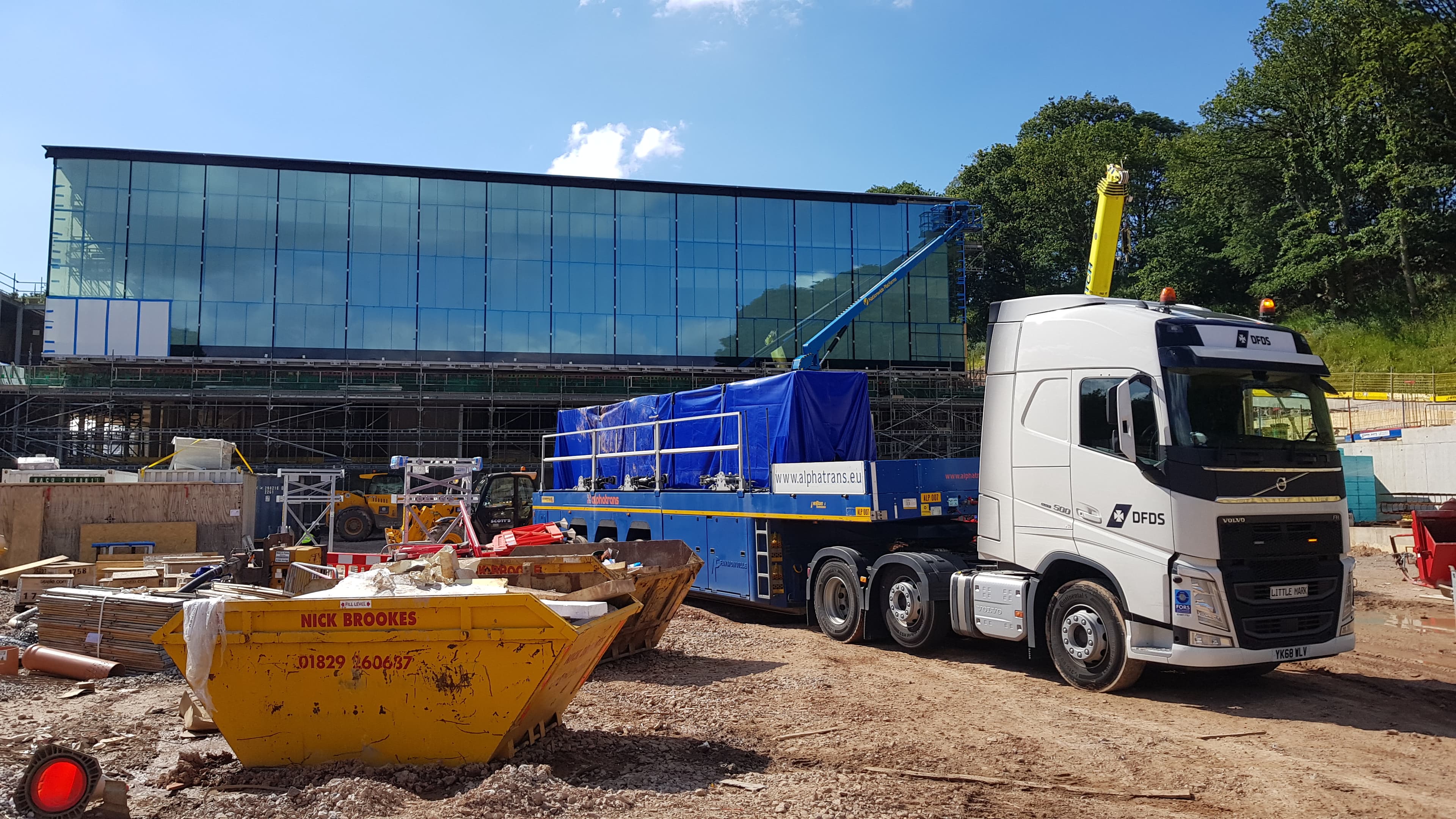 A DFDS truck carrying cargo wrapped in a blue plastic cover is stopped next to a glass building under construction