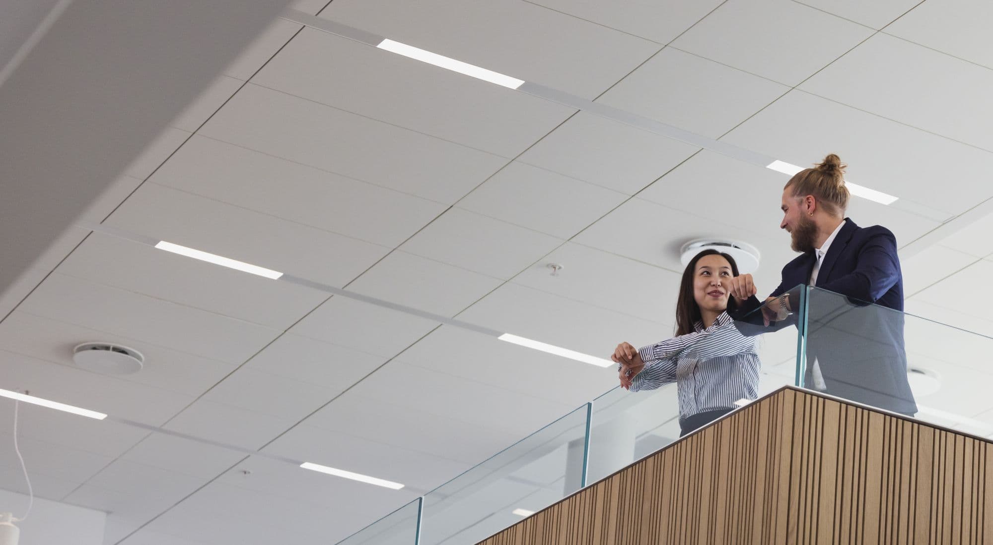 Two office workers, a man & woman, are talking to each other, leaning on the balustrade of a stairs platform in an office