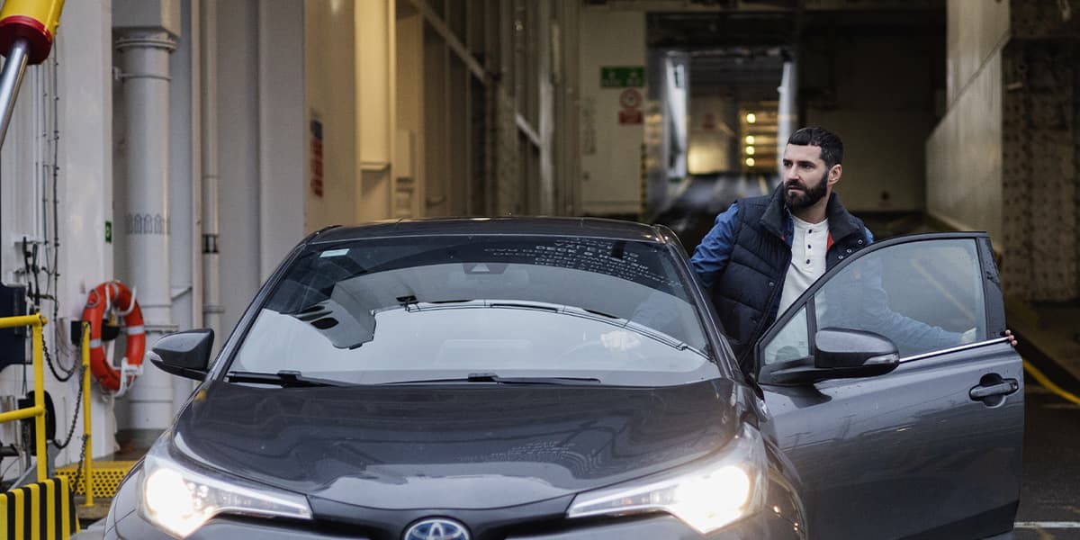 A man in a navy vest stands next to a dark gray Toyota car with the door open, in a parking structure on DFDS ferry.