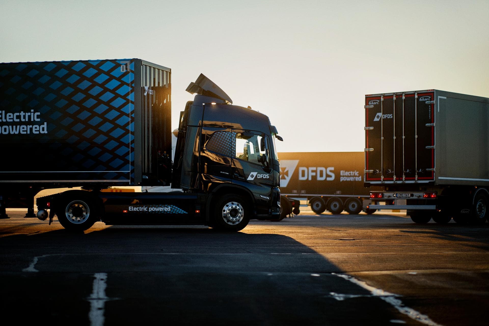 Three electric trucks in dark blue DFDS livery, standing in a circle on a parking ground, within the background of the sunset