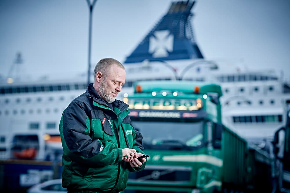 DFDS Logistics truck driver stands in front of a green truck while looking at a mobile phone